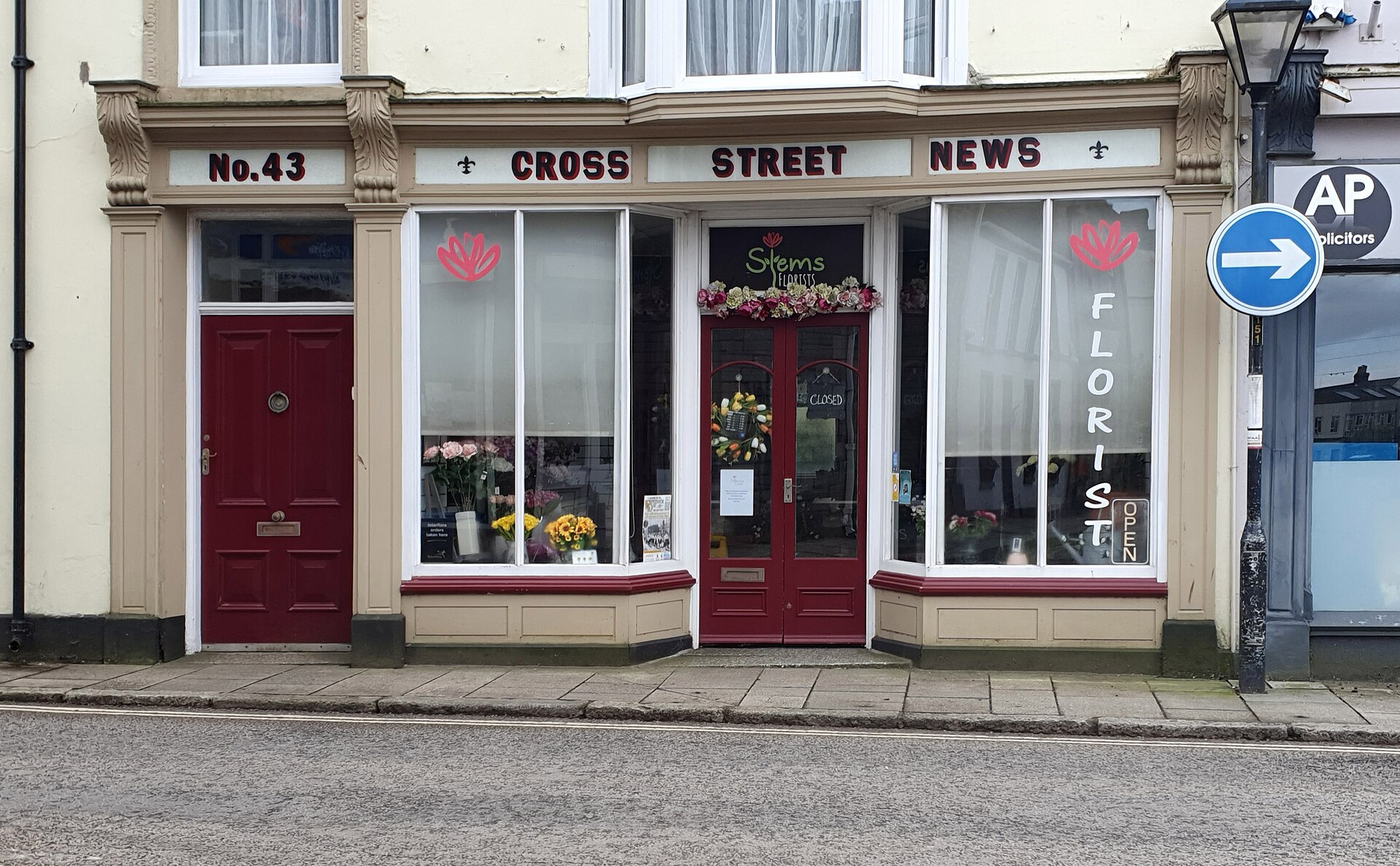 A florist’s shopfront at No. 43 Cross Street, Camborne, Cornwall, with flowers in the bay windows and a dark red door.