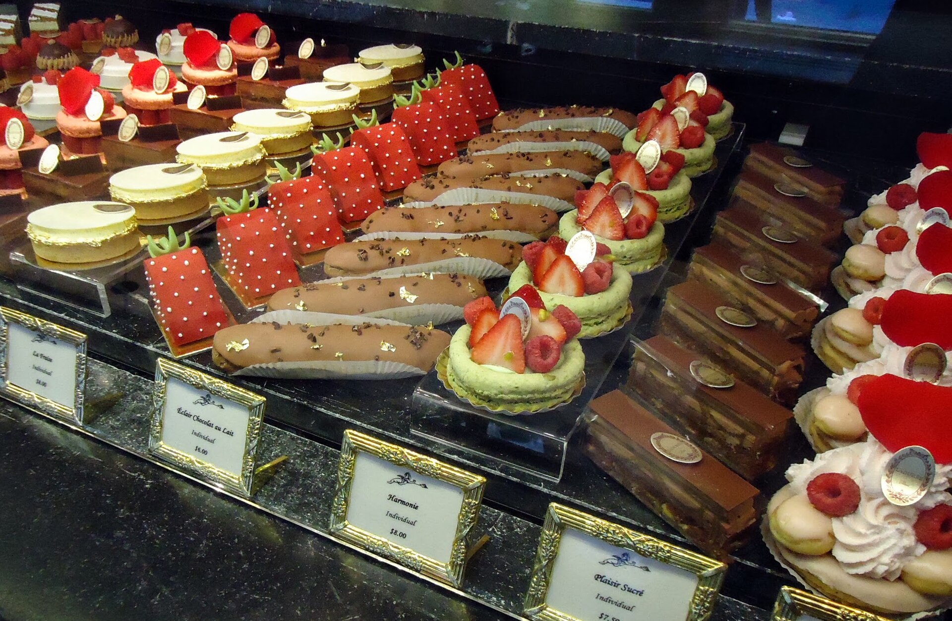 A shop window of pastries and cakes, each on a small label with its price, on a dark marble counter.